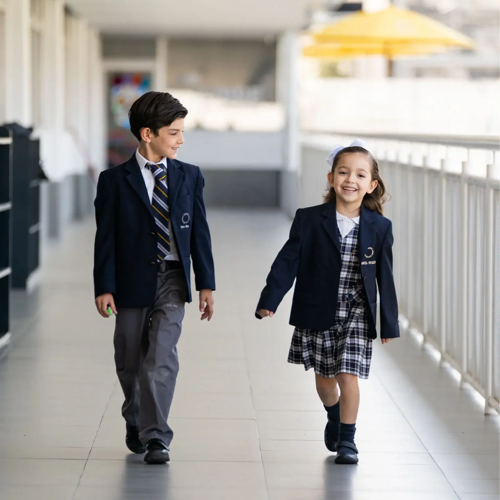Dos estudiantes con uniforme escolar caminan por el pasillo de la escuela sonriendo durante el día.
